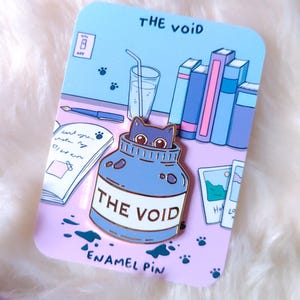 Op de afbeelding: Een blauwe emaille pin met een kat die uit een pot komt met het opschrift "THE VOID". De pin zit op een kaart met illustraties van boeken, een drankje en pootafdrukken. De tekst "ENAMEL PIN" is ook zichtbaar.