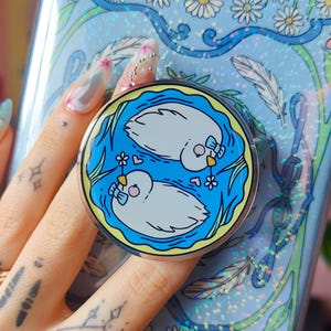 May include: A light blue phone case featuring a pattern of white daisies, green vines, and feathers with a holographic finish. Attached to the centre is a circular phone grip displaying two white birds with blue bows swimming in blue water.