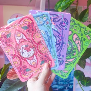 Può includere: Quattro stampe artistiche rettangolari con linee complesse e dettagli olografici. Le carte presentano vivaci schemi di colori rosa, blu, viola e verde con motivi floreali, disegni a spirale e un'illustrazione centrale di un gatto.