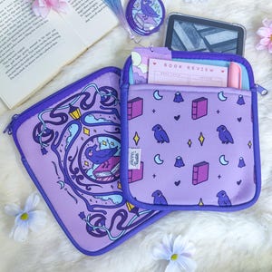 Peut inclure: Deux housses de protection violettes pour livres ou tablettes. L'une présente une illustration circulaire d'un corbeau sur un livre avec des vignes. L'autre a un motif répétitif de corbeaux, livres, lunes et étoiles, contenant une fiche de lecture.