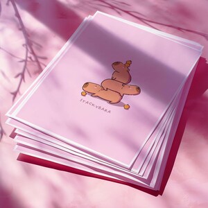May include: A stack of pink art prints with a cartoon illustration of capybaras. The capybaras are stacked, with one balancing a small orange on its head. The text "STACKYBARA" is printed at the bottom of the prints.