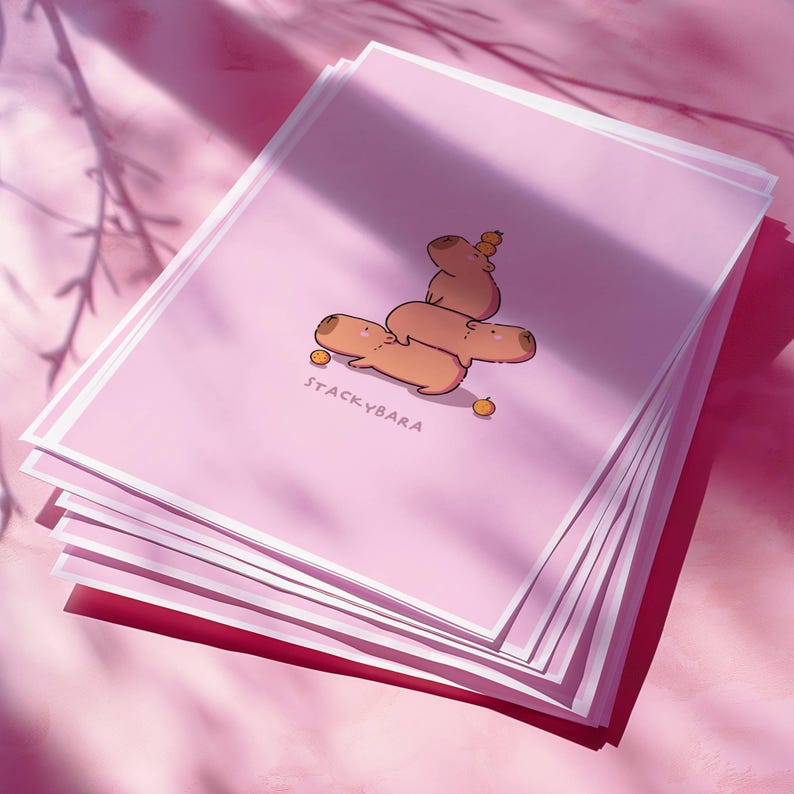 May include: A stack of pink art prints with a cartoon illustration of capybaras. The capybaras are stacked, with one balancing a small orange on its head. The text "STACKYBARA" is printed at the bottom of the prints.