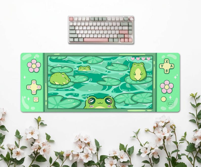 May include: A green and white keyboard rests above a green desk mat. The desk mat features a cartoon frog pond scene with lily pads and frogs. The mat has a game controller design with flower and button details.
