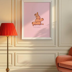 May include: A framed art print showing a stack of cartoon capybaras on a pink backdrop. The capybaras are stacked, with a small carrot on top. The text "STACKYBARA" is printed below the capybaras.