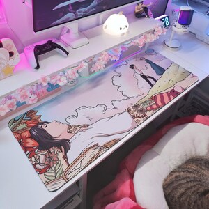 May include: A rectangular desk mat featuring an anime-style illustration of a person lying down, surrounded by flowers and books. The mat has a pastel color scheme with a mountain and cloud scene. A controller and monitor are visible in the background.