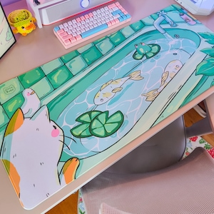 Op de afbeelding: Een rechthoekige bureaumat met een illustratie van een badkamerscène met een kat, vissen en een kikker. De mat heeft een lichtblauw en groen kleurenschema met een witte rand. Een toetsenbord en andere bureau-accessoires zijn zichtbaar.