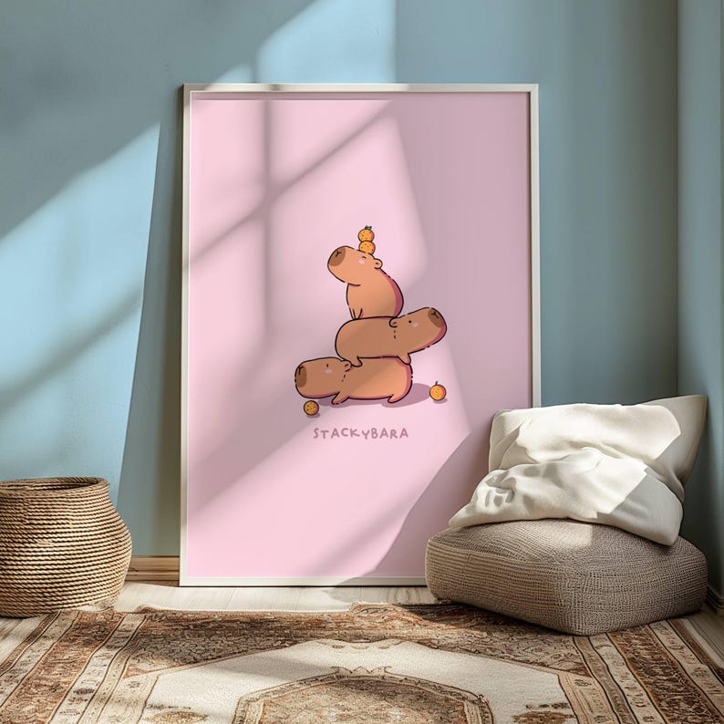 May include: A framed art print with a cartoon illustration of three capybaras balancing on top of each other, with small orange fruits. The print has a pink background and the text "STACKYBARA" at the bottom.