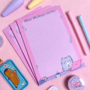 Peut inclure: Bloc-notes rose avec un motif quadrillé et le texte "VERY SERIOUS NOTES". Le bloc-notes présente un chat de dessin animé travaillant sur un ordinateur portable. D'autres articles de papeterie sont présents.