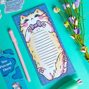 May include: A notepad with a cartoon cat illustration, featuring a light yellow and pink cat with a purple bow tie, on a blue grid background. A pink pen and a bookmark that says "You Pawsed Here" are also in the image.