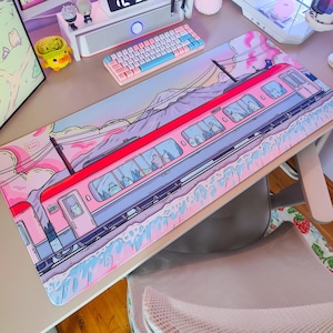 Puede incluir: Una alfombrilla de escritorio rosa y azul con un tren de dibujos animados que viaja por un paisaje montañoso. El tren tiene un techo rojo y laterales azules, con ventanas que muestran pasajeros. El fondo es un cielo pastel.