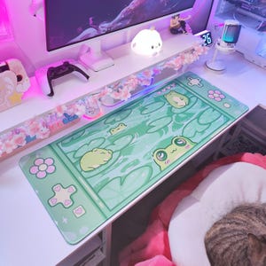 May include: A rectangular green mousepad featuring a cartoon frog design. The mousepad has a light green background with lily pads and frogs. The border of the mousepad has flower and heart shapes.