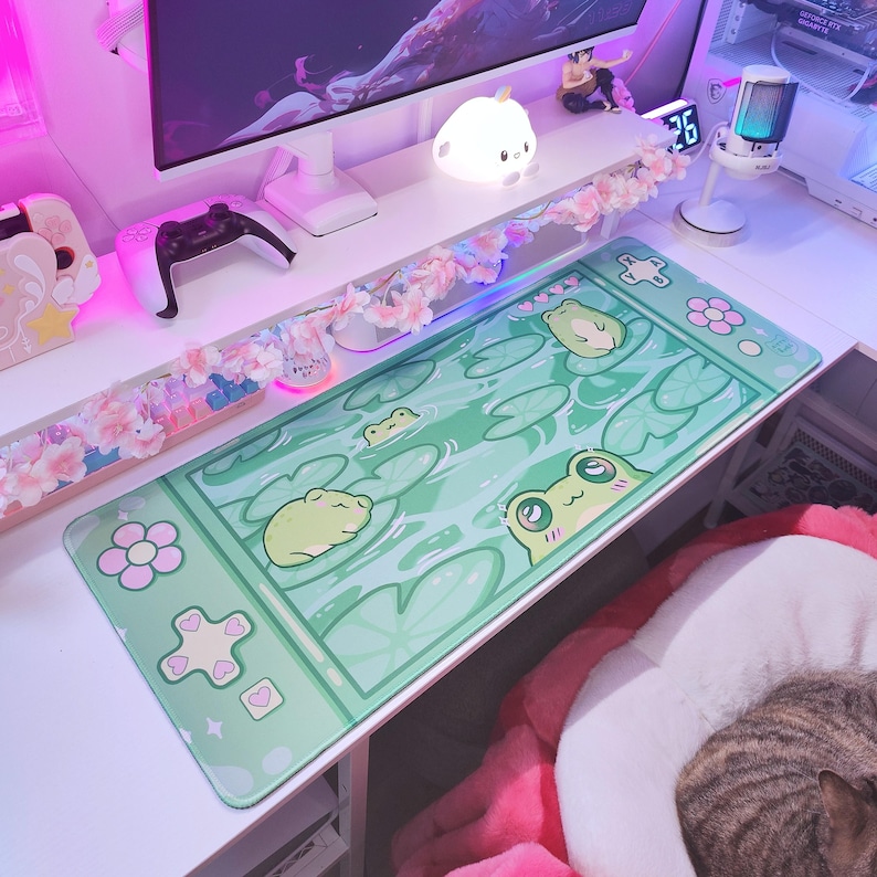 May include: A rectangular green mousepad featuring a cartoon frog design. The mousepad has a light green background with lily pads and frogs. The border of the mousepad has flower and heart shapes.