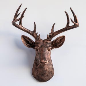 May include: A bronze-colored deer head wall mount with detailed antlers and textured fur. The sculpture is mounted on a white wall, showcasing the intricate design and rustic aesthetic. The deer head is a decorative item.
