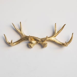 May include: A gold-colored antler wall decor piece. The antlers are mounted horizontally and have multiple tines. The piece is likely made of metal and is designed to be mounted on a wall. The overall design is rustic and decorative.