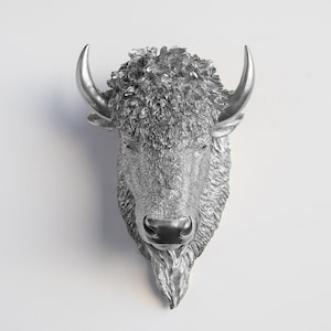 Puede incluir: Una escultura de cabeza de bisonte plateada con detalles de pelaje y cuernos curvados. La escultura está montada sobre un fondo blanco, mostrando la intrincada textura y el diseño de los rasgos del animal. Esta pieza decorativa añade un toque de encanto rústico.