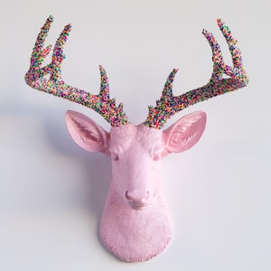 May include: A pink deer head wall mount with colourful, textured antlers. The deer head is a solid, matte pink colour, and the antlers are covered in small, multicoloured beads. The deer head is mounted on a white wall.