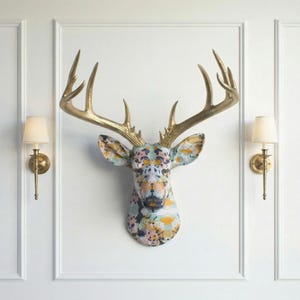 May include: A decorative deer head with gold antlers and a patterned face in various colours. The head is mounted on a white wall, with two wall-mounted lamps either side. The antlers provide a striking contrast to the patterned face.
