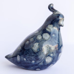 May include: A ceramic bird sculpture in shades of blue with white and grey speckled patterns. The bird has a curved beak and a small, curved crest. The sculpture is glossy and appears to be handmade.