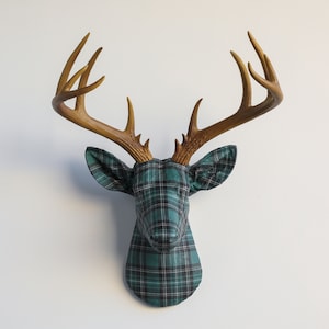 May include: A decorative deer head with brown antlers and a head covered in a green and black plaid fabric. The deer head is mounted on a white wall, creating a rustic and stylish wall decor piece.