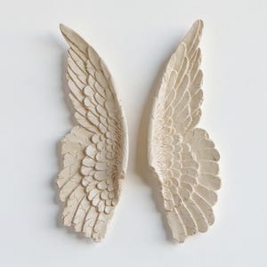 Peut inclure: Deux ailes d'ange textur&eacute;es, de couleur blanc cass&eacute;, sur fond blanc. Les ailes pr&eacute;sentent des motifs de plumes d&eacute;taill&eacute;s et un aspect l&eacute;g&egrave;rement vieilli, sugg&eacute;rant un style vintage ou antique. Il s'agit probablement d'objets d&eacute;coratifs muraux.