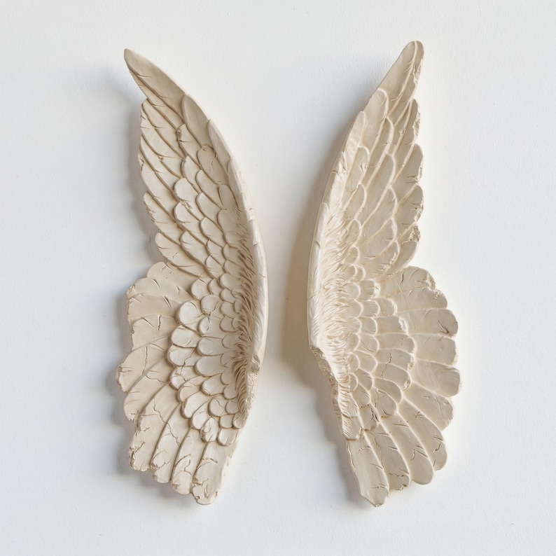Peut inclure: Deux ailes d'ange textur&eacute;es, de couleur blanc cass&eacute;, sur fond blanc. Les ailes pr&eacute;sentent des motifs de plumes d&eacute;taill&eacute;s et un aspect l&eacute;g&egrave;rement vieilli, sugg&eacute;rant un style vintage ou antique. Il s'agit probablement d'objets d&eacute;coratifs muraux.