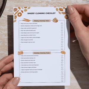 May include: A person holding a white "Bakery Cleaning Checklist" with decorative baked goods. The checklist has two sections: "Weekly Cleaning Tasks" and "Monthly Cleaning Tasks". A silver pen and a glass of liquid are also visible.