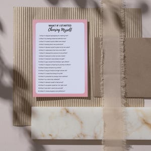 Op de afbeelding: Een roze en witte kaart met de tekst "WHAT IF I STARTED CHOOSING MYSELF" en een lijst met vragen. De kaart ligt op een getextureerd beige oppervlak met andere decoratieve elementen, waaronder een lint en een marmerachtig blok.