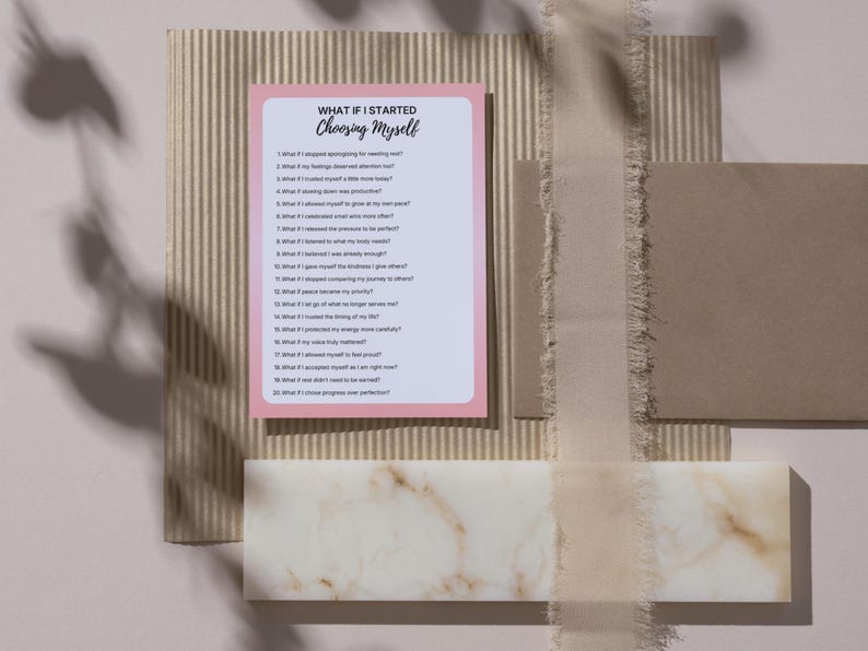 Op de afbeelding: Een roze en witte kaart met de tekst "WHAT IF I STARTED CHOOSING MYSELF" en een lijst met vragen. De kaart ligt op een getextureerd beige oppervlak met andere decoratieve elementen, waaronder een lint en een marmerachtig blok.