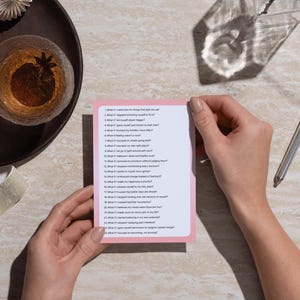 Op de afbeelding: Een persoon houdt een roze kaart met een lijst met vragen vast. Een glas amberkleurige vloeistof met steranijs staat op een donkere houten schaal. Een zilveren pen ligt op een licht oppervlak. De tekst op de kaart nodigt uit tot zelfreflectie.