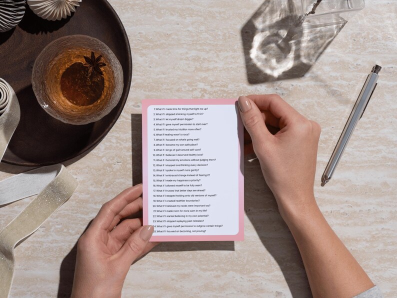 Op de afbeelding: Een persoon houdt een roze kaart met een lijst met vragen vast. Een glas amberkleurige vloeistof met steranijs staat op een donkere houten schaal. Een zilveren pen ligt op een licht oppervlak. De tekst op de kaart nodigt uit tot zelfreflectie.