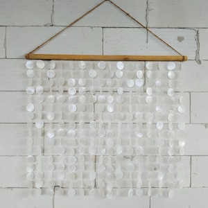 Peut inclure: Une suspension murale composée de multiples brins verticaux de disques de coquillages blancs irisés, suspendus à une tige en bois horizontale. Une fine cordelette en ficelle brune est fixée aux deux extrémités de la tige pour la suspension.
