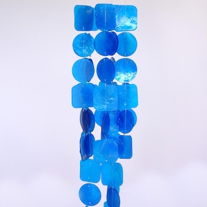 May include: A hanging wind chime with a wooden top and strings of blue, square, and circular shapes. The shapes are made of a translucent material, creating a shimmering effect. The wind chime is suspended against a white background.