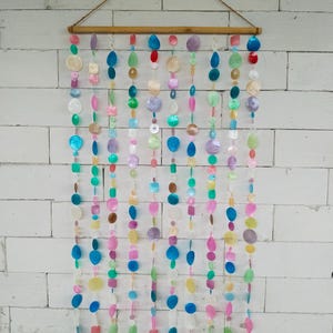 Peut inclure: Une suspension murale composée de multiples brins verticaux de disques de coquillages irisés multicolores suspendus à une tige en bois horizontale. Les pièces de coquillages circulaires et ovales présentent une variété de teintes vives, notamment bleu, rose, vert, violet et blanc, créant un effet de cascade superposé sur toute la longueur des brins.