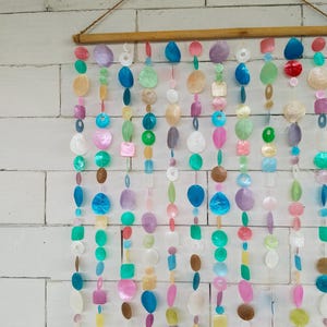 Puede incluir: Una decoración de pared colgante que presenta múltiples hilos verticales de discos de concha capiz iridiscentes y formas geométricas multicolores, suspendidos de una varilla de madera horizontal con un cordel marrón.