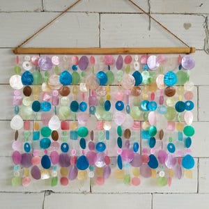 Puede incluir: Un colgante de pared compuesto por múltiples hilos verticales de discos de concha iridiscentes, circulares y ovalados, en tonos azul, verde, rosa y blanco. Los hilos cuelgan de una varilla de madera horizontal con un cordón marrón fino.