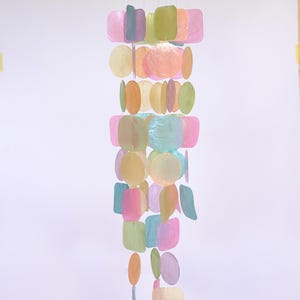 Op de afbeelding: Een kleurrijke windgong met een houten bovenkant en een waterval van iriserende, pastelkleurige schelpstukken. De vormen omvatten vierkanten, cirkels en druppels in tinten roze, groen, geel en blauw. Decoratief.