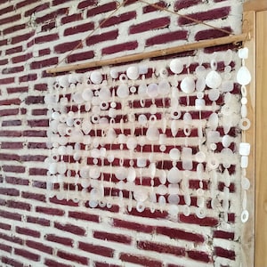 Puede incluir: Un tapiz compuesto por múltiples hilos verticales de discos de conchas blancas translúcidas y formas geométricas suspendidos de una varilla de bambú horizontal con un fino lazo de cuerda marrón para colgar en la pared.