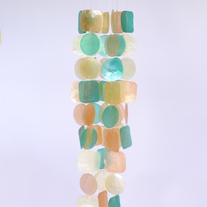 Peut inclure: Un carillon décoratif avec un dessus en bois et une cascade de morceaux de coquillages irisés et colorés. Les coquillages sont de formes variées, dont des carrés et des cercles, et dans des tons de vert, de jaune et de pêche.