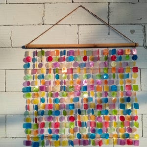 May include: A wall hanging composed of numerous translucent, multi-coloured shell discs in circular and square shapes. The discs are arranged in vertical strands suspended from a horizontal wooden rod with a thin brown string for hanging.