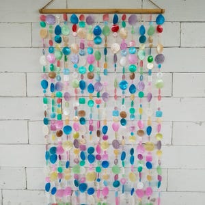 Puede incluir: Un tapiz decorativo con múltiples hilos verticales de discos de concha iridiscentes y multicolores en tonos azul, rosa, verde, morado y blanco. Los hilos cuelgan de una varilla de madera horizontal con un cordel marrón para colgarlo.