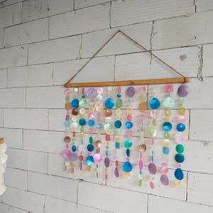 May include: A wall hanging featuring multiple vertical strands of circular and oval-shaped iridescent shell discs in shades of blue, pink, purple, green, and white. The strands are suspended from a horizontal wooden rod with a brown twine hanger.