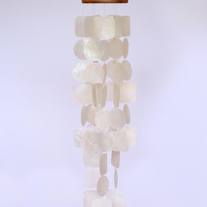 May include: A hanging wind chime with a wooden top and strands of white, square, and circular shell pieces. The shell pieces are arranged in a cascading pattern, creating a decorative and airy design. The wind chime is suspended against a plain background.