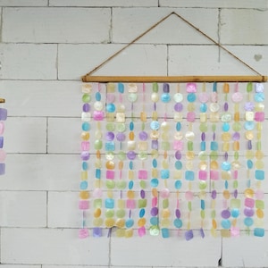 Puede incluir: Un tapiz de pared y un móvil más pequeño hechos de discos de concha iridiscentes y multicolores en tonos rosa, azul, morado, amarillo y verde. El tapiz presenta filas de estos discos suspendidos de una varilla de madera con un cordel.