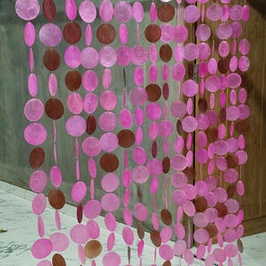Peut inclure: Un rideau séparateur de pièce suspendu composé de multiples brins verticaux de disques de coquillages circulaires, plats, translucides, roses et bruns. Les disques sont enfilés sur de fins filaments transparents et suspendus à une tringle en bois.