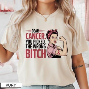 Puede incluir: Camiseta de color marfil con el texto "DEAR CANCER, YOU PICKED, THE WRONG BITCH" en rojo y blanco. La camiseta presenta un gráfico de una mujer flexionando su brazo, con una camisa rosa y una diadema de lunares rojos. Una cinta rosa está en su brazo.