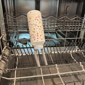 Pode incluir: Uma garrafa para bebé cilíndrica com um padrão floral de flores rosa, amarelas e azuis sobre um fundo branco. A garrafa está colocada numa máquina de lavar louça.