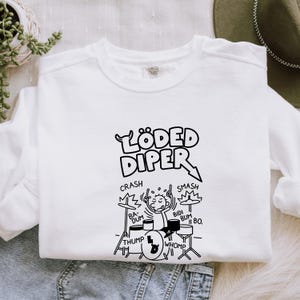 May include: White sweatshirt with a black graphic of a drummer playing a drum set. The text "LÖDED DIPER" is above the drummer, with drum sounds like "CRASH", "SMASH", "BA-DUM", "BIDI BUM", "THUMP", and "WHOMP" around the drum set.