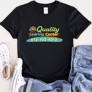 May include: Black t-shirt with the words "Quality Learning Center" in green and yellow, along with a phone number. The shirt is paired with white sneakers and blue jeans.