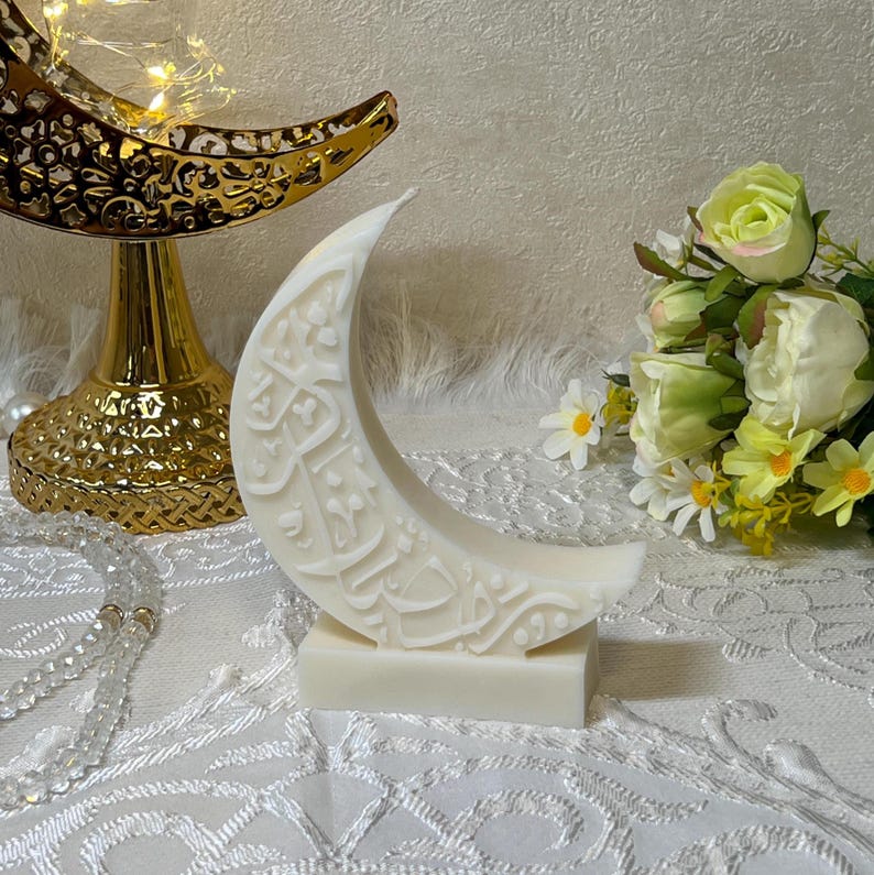 Stilvolle weiße Halbmondkerze mit feiner arabischer Schrift, präsentiert auf einer glatten Marmorplatte. Umgeben von goldenen Accessoires, floralen Akzenten und sanfter Beleuchtung entsteht eine warme, einladende Ramadan-Atmosphäre. Die Kombination aus moderner Form und traditioneller Kalligrafie macht diese Kerze zu einem dekorativen Highlight für Wohnräume, Festtafeln oder als besonderes Geschenk.
