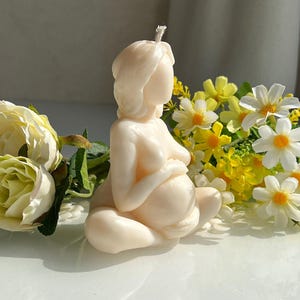 Könnte beinhalten: Eine cremefarbene Kerze in Form einer schwangeren Person, die sitzt und die Hände sanft auf dem Bauch hält. Die Kerze hat einen Docht oben und ist von gelben und weißen Blumen umgeben.